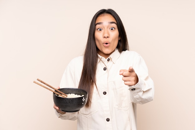 Joven india aislada en beige sorprendido y apuntando hacia el frente mientras sostiene un plato de fideos con palillos