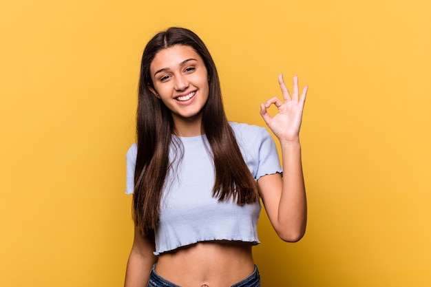 Joven india aislada en la pared amarilla guiña un ojo y tiene un gesto bien con la mano.