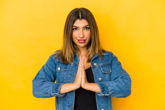 Joven india aislada en la pared amarilla rezando, mostrando devoción, persona religiosa en busca de inspiración divina.