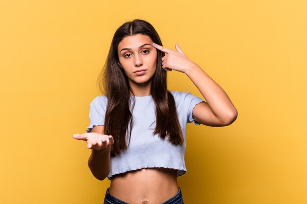 Joven india aislada en la pared amarilla sosteniendo y mostrando un producto en la mano