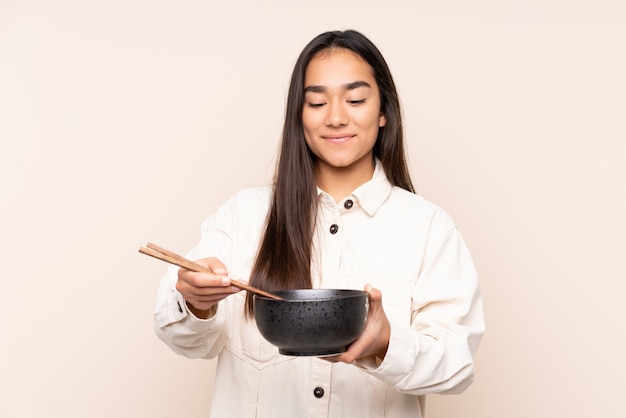 Joven india aislada en la pared de color beige con un tazón de fideos con palillos