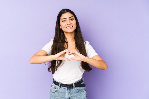 Joven india aislada en la pared púrpura sonriendo y mostrando una forma de corazón con las manos.