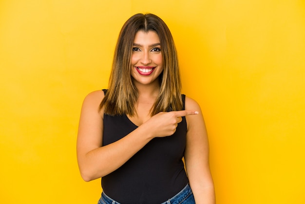 Joven india aislada sobre fondo amarillo sonriendo y apuntando a un lado, mostrando algo en el espacio en blanco.