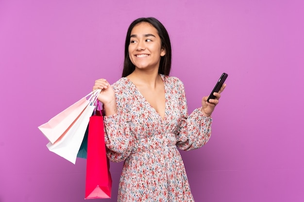 Joven India aislada sobre fondo púrpura sosteniendo bolsas de la compra y un teléfono móvil