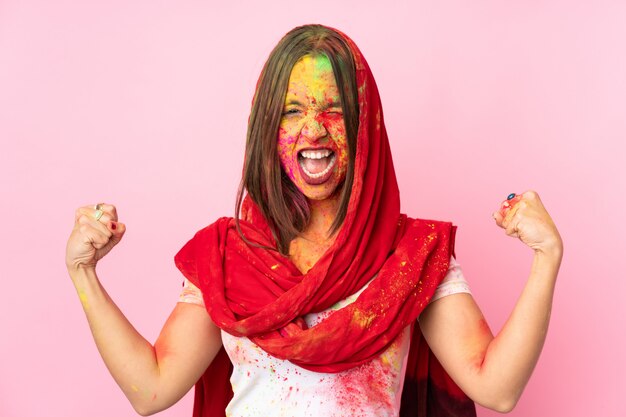 Joven india con coloridos polvos de holi en la cara en la pared de color rosa haciendo un gesto fuerte