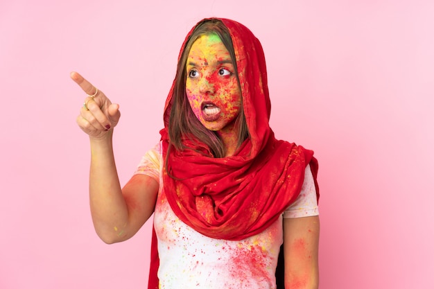 Joven india con coloridos polvos de holi en la cara en la pared rosa apuntando lejos