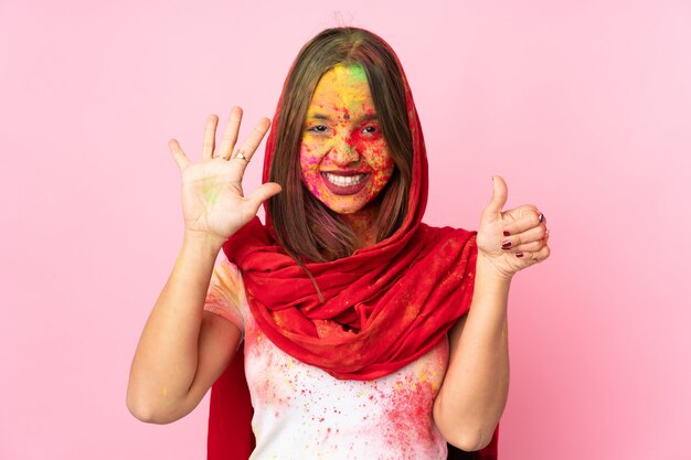 Joven india con coloridos polvos de holi en la cara en la pared rosa contando seis con los dedos