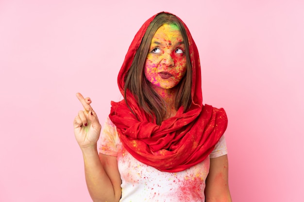 Joven india con coloridos polvos de holi en la cara en la pared rosa con los dedos cruzados y deseando lo mejor