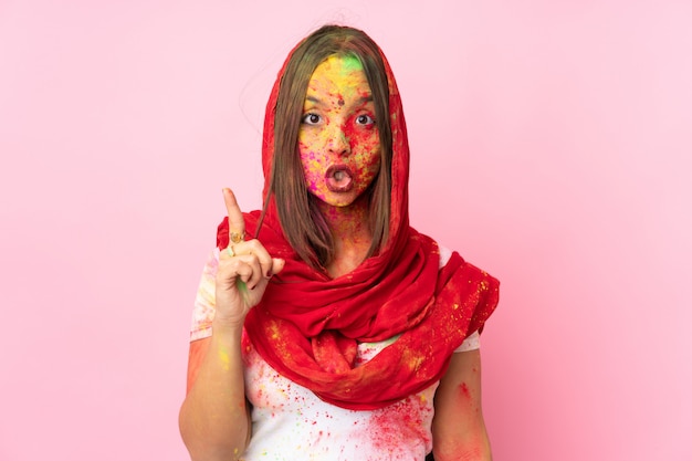 Joven india con coloridos polvos de holi en la cara en la pared rosa con la intención de darse cuenta de la solución mientras levanta un dedo