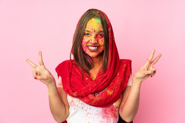 Joven india con coloridos polvos de holi en la cara en la pared rosa que muestra el signo de la victoria con ambas manos