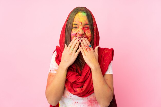 Joven india con coloridos polvos de holi en la cara en la pared rosada feliz y sonriente cubriendo la boca con las manos