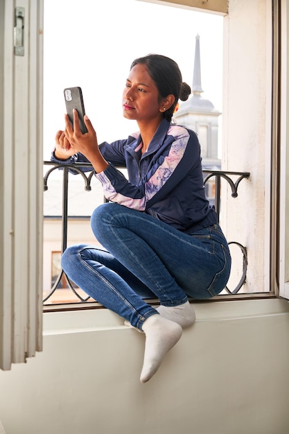 Una joven india conectada y entretenida disfruta de su teléfono mientras se sienta en el alféizar de la ventana