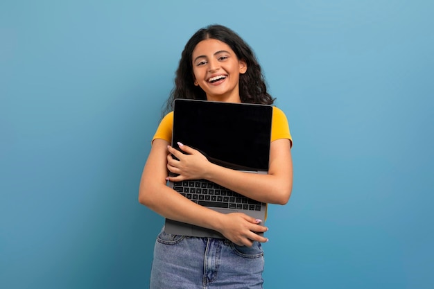 Una joven india encantada abrazando una computadora portátil con pantalla en blanco