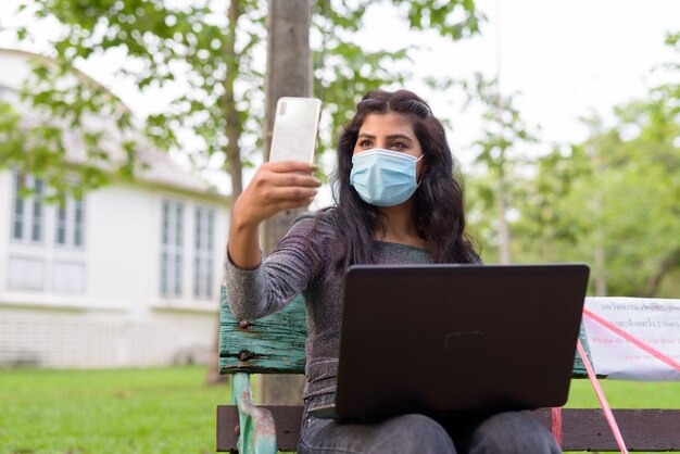 Joven india con máscara de videollamadas mediante teléfono mientras está sentado con distancia en un banco del parque