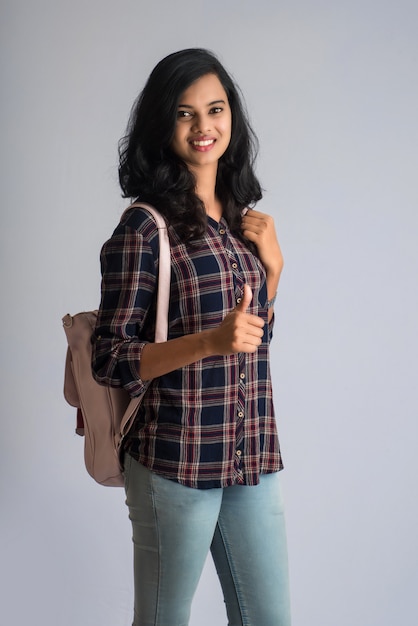 Joven india con mochila mostrando el pulgar y posando sobre una pared gris