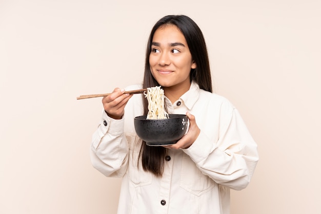 Joven india en pared beige sosteniendo un tazón de fideos con palillos