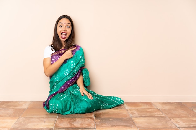 Joven india sentada en el suelo sorprendida y apuntando hacia el lado