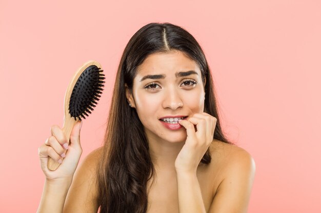 Joven india sosteniendo un cepillo para el pelo mordiendo las uñas, nervioso y muy ansioso.