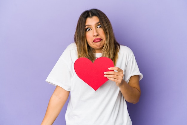 Joven india sosteniendo un corazón de San Valentín aislado se encoge de hombros y abre los ojos confundidos.