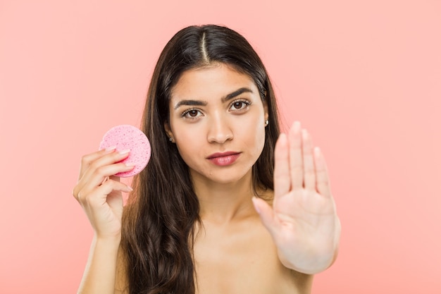 Joven india sosteniendo un disco de cuidado de la piel facial de pie con la mano extendida mostrando la señal de stop, previniéndote.