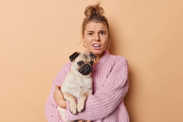 Una joven indignada y descontenta frunce el ceño y mira descontenta a la cámara escucha malas noticias del veterinario sobre la enfermedad de las mascotas sostiene poses de perro pug pedigrí contra un fondo beige siendo aficionado a los animales