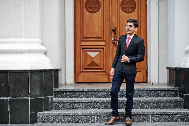 Un joven indio con anteojos se viste con traje negro y corbata roja al aire libre contra la puerta del edificio