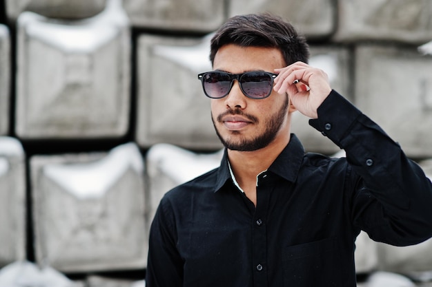 Un joven indio casual con camisa negra y gafas de sol posó contra bloques de piedra