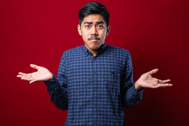 Joven indio guapo con camisa casual sobre fondo rojo expresión desorientada y confusa con los brazos y las manos levantadas. Concepto de duda.