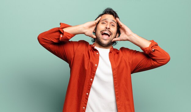 Joven indio guapo levantando las manos a la cabeza, con la boca abierta, sintiéndose extremadamente afortunado, sorprendido, emocionado y feliz