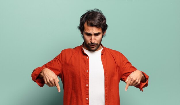 Joven indio guapo sintiéndose conmocionado, con la boca abierta y asombrado, mirando y apuntando hacia abajo con incredulidad y sorpresa.