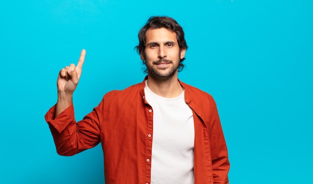 Joven indio guapo sonriendo alegre y felizmente, apuntando hacia arriba con una mano para copiar el espacio