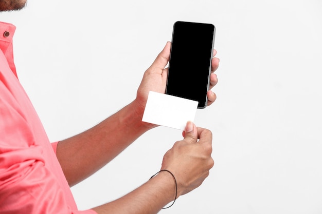 Joven indio mostrando la tarjeta y el teléfono inteligente en la pared blanca