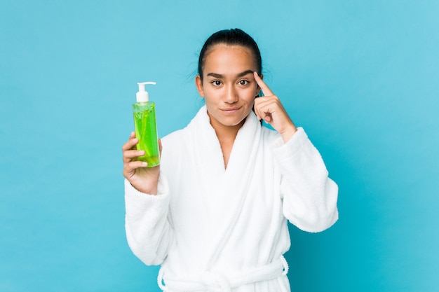 Joven indio de raza mixta sosteniendo una botella de aloe vera apuntando su sien con el dedo, pensando, centrado en la tarea.