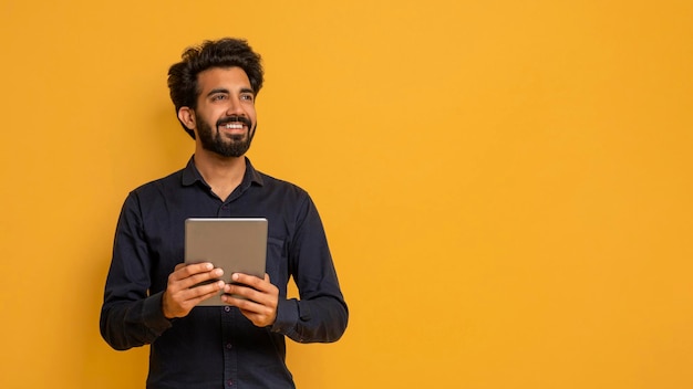 Joven indio sosteniendo una tableta digital y mirando hacia otro lado pensativo soñador hombre oriental con un dispositivo moderno en las manos de pie sobre el fondo amarillo del estudio pensando en una buena oferta panorámica