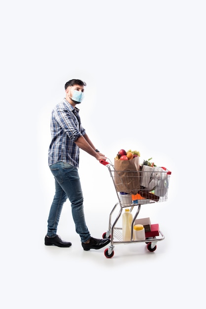 Joven indio usa mascarilla con carrito de compras o carrito lleno de comestibles, verduras y frutas. Foto de cuerpo entero aislado sobre pared blanca