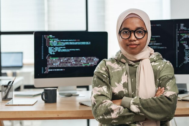 Joven ingeniera de ti confiada en hijab mirando a la cámara mientras está sentada en el lugar de trabajo contra com