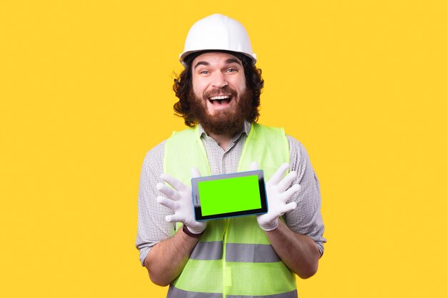 Un joven ingeniero alegre sostiene una tableta nueva mirando y sonriendo a la cámara cerca de una pared amarilla