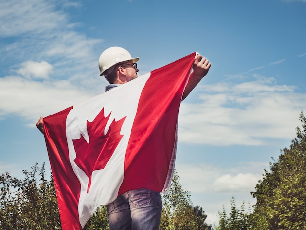 Joven ingeniero, casco blanco y bandera canadiense en el parque contra el telón de fondo de árboles verdes y la puesta de sol, mirando a lo lejos. De cerca. Concepto de trabajo y empleo.