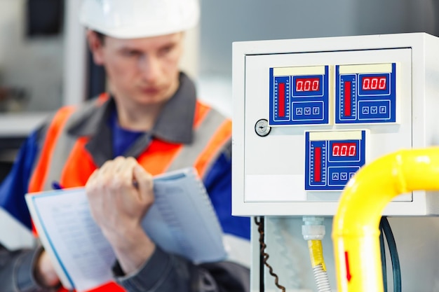 Foto joven ingeniero a casco y registros de chaleco protector en lecturas de registro de dispositivos o consumo de gas en sala de calderas