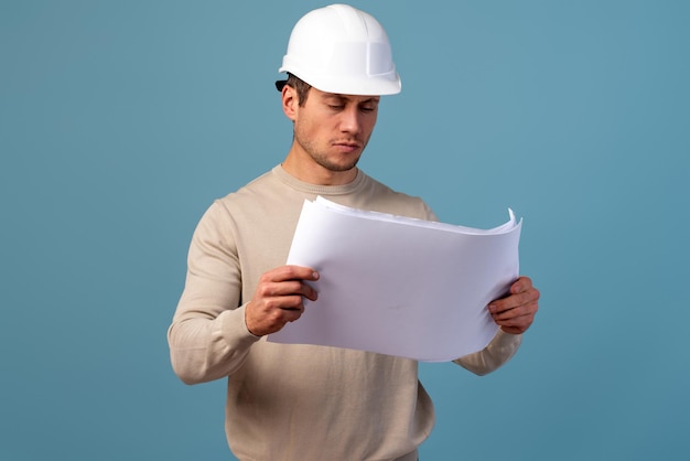 Joven ingeniero civil apuesto sosteniendo un plano de papeleo y mirándolo mientras está de pie en el estudio. trabajador, llevando, sombrero duro, en, aislado, fondo azul