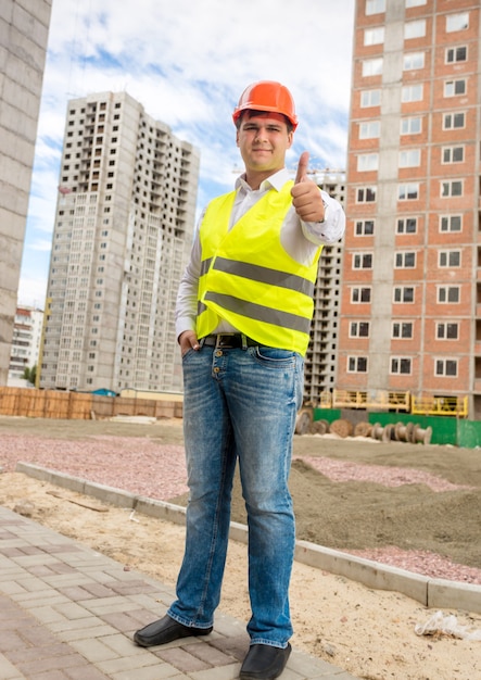 Joven ingeniero de construcción sonriente posando en el sitio de construcción con el pulgar hacia arriba