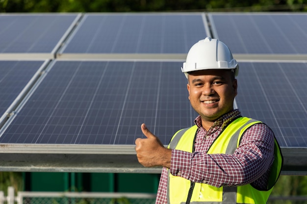 Joven ingeniero eléctrico asiático parado frente a la granja de paneles de células solares. Él muestra los pulgares hacia arriba. Concepto de energía del generador solar.