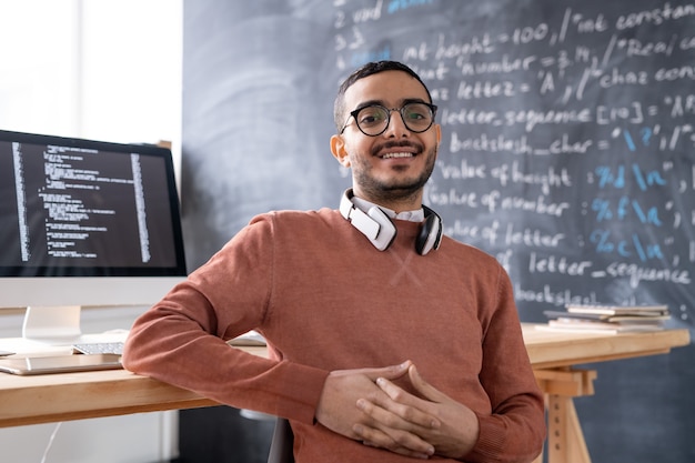 Joven ingeniero informático de contenido en anteojos sentado contra la mesa con monitor de computadora y pizarra