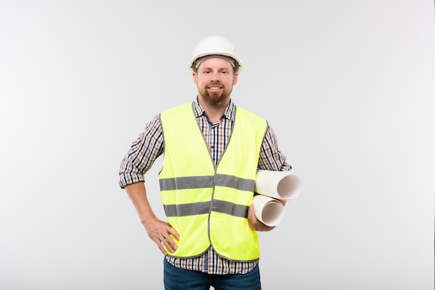 Joven ingeniero o capataz alegre en casco protector y ropa de trabajo sosteniendo planos enrollados de forma aislada