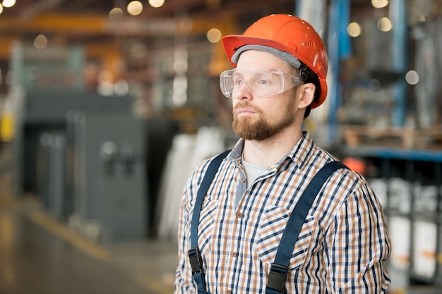 Joven ingeniero serio en casco, anteojos y ropa de trabajo dentro de una gran fábrica moderna