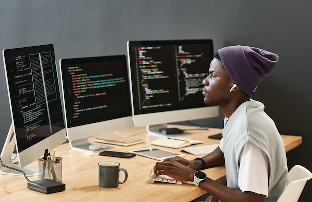 Joven ingeniero de ti serio en ropa casual mirando datos codificados en pantallas de computadora mientras escribe en ke