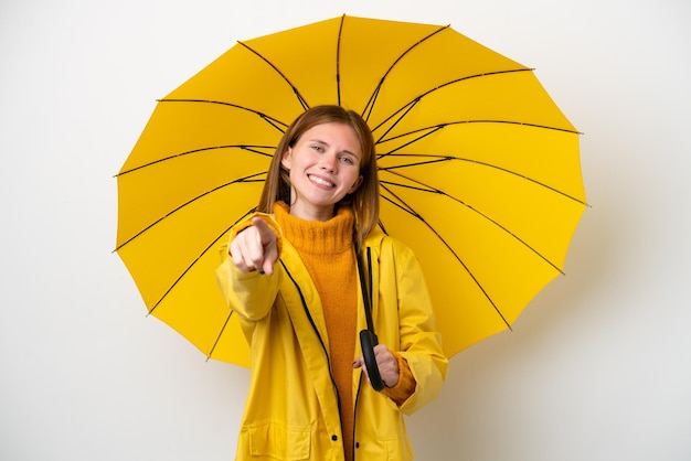 Joven inglesa con abrigo impermeable y paraguas aislado de fondo blanco apuntando al frente con expresión feliz