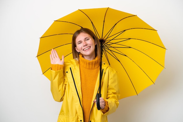 Joven inglesa con abrigo impermeable y paraguas aislado de fondo blanco saludando con la mano con expresión feliz