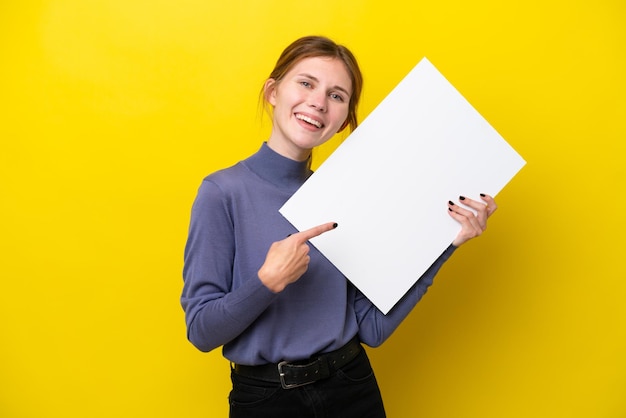 Joven inglesa aislada de fondo amarillo sosteniendo un cartel vacío con expresión feliz y apuntándolo