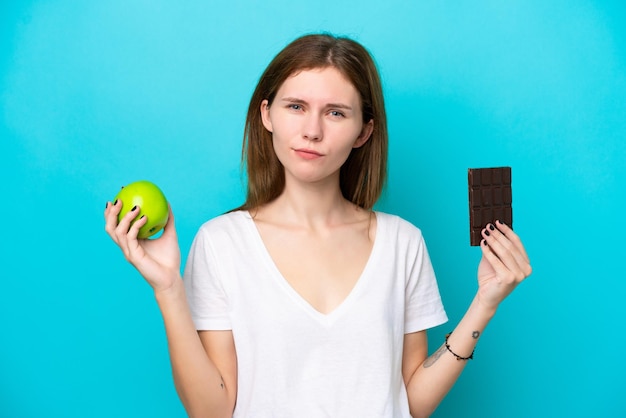 Joven inglesa aislada de fondo azul que tiene dudas mientras toma una tableta de chocolate en una mano y una manzana en la otra